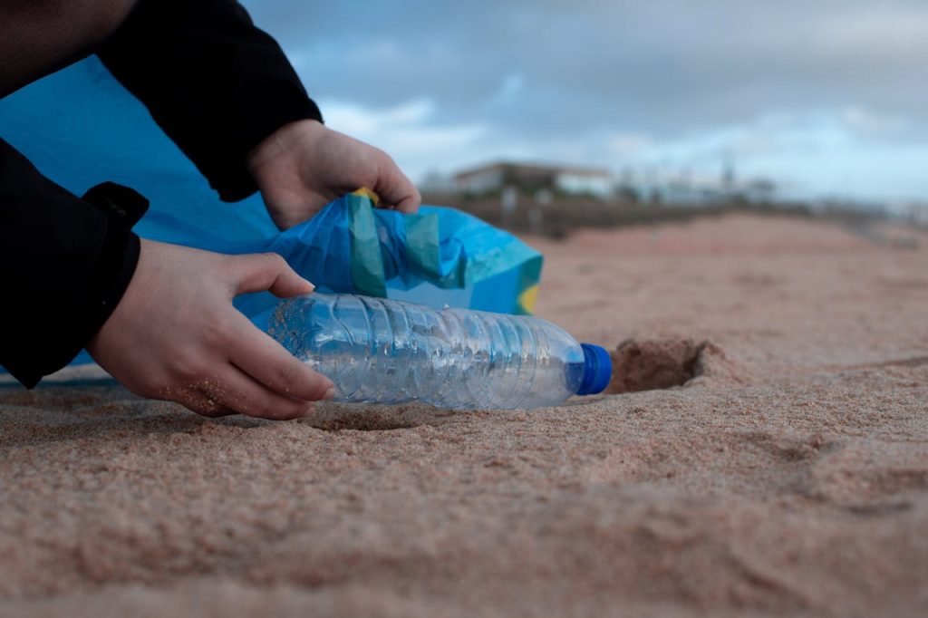 Hands collecting a plastic water bottle from the sandy beach, promoting environmental awareness.