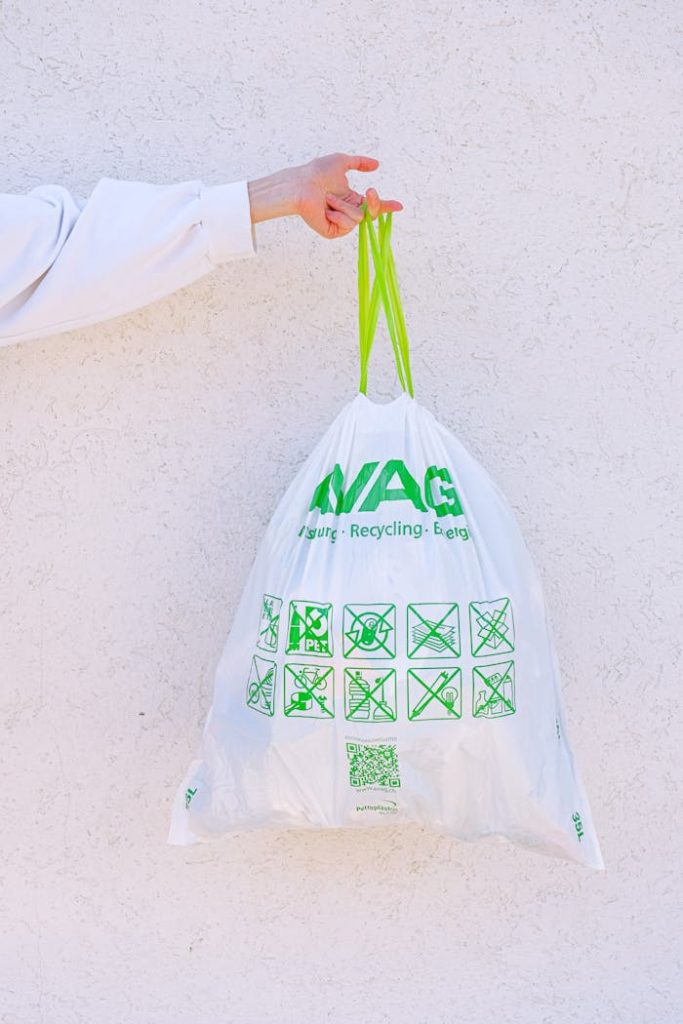 A person holding a white recycling bag with green symbols against a white wall, promoting sustainability.